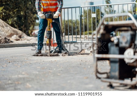 Craftsman in uniform with drill repairing asphalt during roadwork