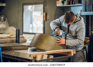 A craftsman is upholstering a leather chair at the furniture workshop. A focused artisan is upholstering a chair with eco-leather material and making handmade furniture at his workshop.