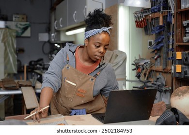 Craftswoman with a headband seen working in workshop using laptop while shaping wooden piece on workbench. Tools and equipment seen neatly organized in background - Powered by Shutterstock