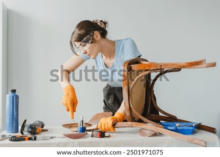 Craftswoman is using a chisel to restore an old wooden chair in her workshop, surrounded by tools and materials. She is wearing safety glasses and gloves for protection