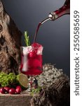 Cranberry cocktail with ice, lime, and rosemary on a old wooden board. Liquor is poured into a glass with ice. North nature background with snags and moss.