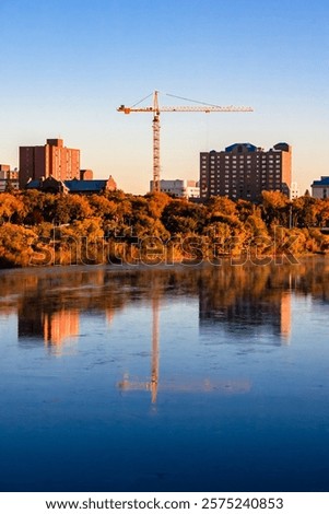 A crane is in the background of a city with a river in the foreground. The sky is blue and the sun is setting