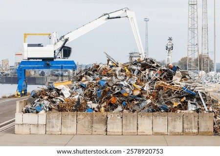 Crane at recycling facility loading large scrap metal pile with wind turbines in background. Industrial area scene waste management and resource reuse concept
