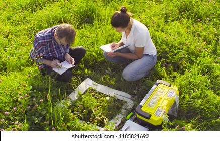 Crane Shot, Top View. Two Ecologist Examining Plants On The Meadow. Woman Writing Something In The Diary. Field Work