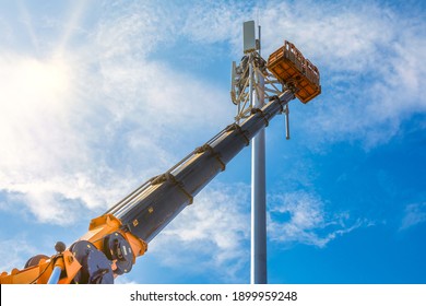 Crane With Telescopic Boom Lift Used As An Aerial Working Platform. Worker Install Cellular Base Station With Transmitters 3G, 4G, And Antennas On Cell Tower On Blue Sky Background.