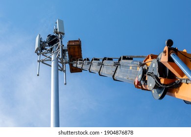 Crane With Telescopic Boom Lift Used As An Aerial Working Platform. Worker Install Cellular Base Station With Transmitters 3G, 4G, And Antennas On Cell Tower On Blue Sky Background. Focus On Tower