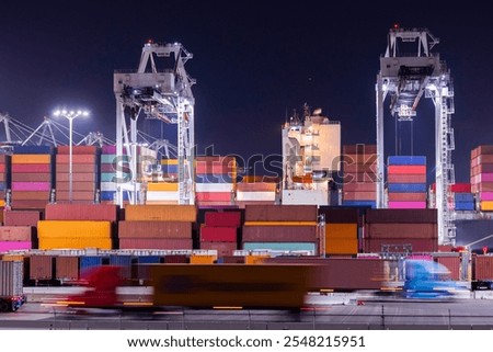 Cranes unload intermodal containers from a ship at port, with motion blurred semi trucks rush in the foreground.