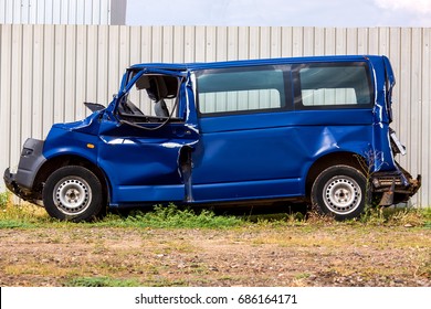 The Crash Car, Broken Minibus After Accident. Failed Blue Van On Country Road, Near A Metal Gray Fence.