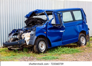 The Crash Car, Broken Minibus After Accident. Failed Blue Van On Country Road, Near A Metal Gray Fence.