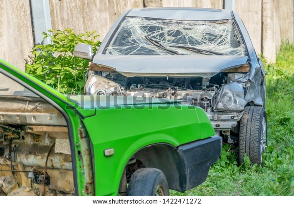 a crashed car after an accident with a broken\
windshield costs near a concrete fence next to another disassembled\
car on a sunny day