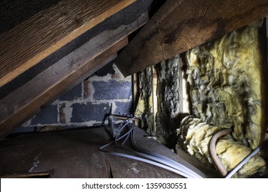 Crawl space under the eves of a house showing old fibreglass insulation, pipework, rafters, breezeblock construction and old boarding. - Shutterstock ID 1359030551