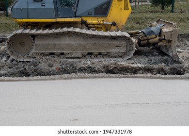 Crawler Bulldozer With Yellow Cab With Exposed Blade And Mud-spattered Tracks Stands On Excavated Soil Surface Of Ground Next To Strip Of Path Rolled Up By Road Roller At Road Works Site.