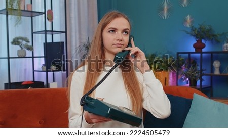 Crazy adult girl in casual shirt talking on wired vintage old-fashioned retro telephone of 80s, fooling, making silly faces. Young woman sitting on orange couch at home living room making conversation