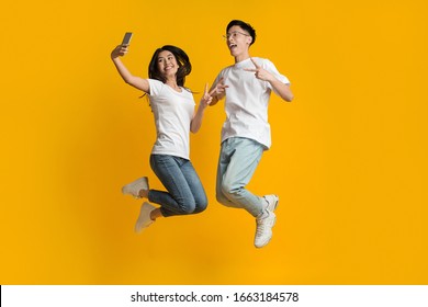 Crazy asian young couple jumping up and taking selfie on smartphone, yellow background, empty space