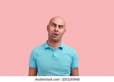 Crazy Bald Unshaven Homosexual Man In Playful Mood Makes Funny Face, Crosses Eyes And Sticks Out Tongue, Plays Fool, Has Fun, Gay Friendly, Wears Blue Polo Shirt, Stands Over Pink Background