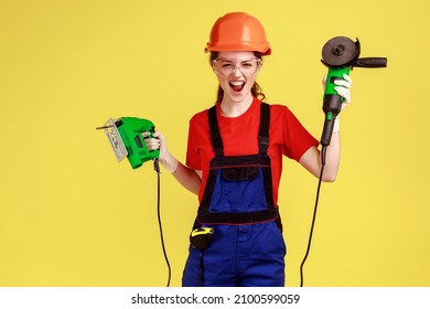 Crazy builder woman holding grinding saw and fretsaw in hands, having crazy facial expression, screaming, wearing overalls and protective helmet. Indoor studio shot isolated on yellow background.