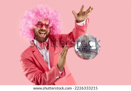 Crazy cheerful and funny man spins little shiny disco ball isolated on pink background. Young Caucasian man in pink wig, glasses and coral suit is having fun and enjoying party. Banner.