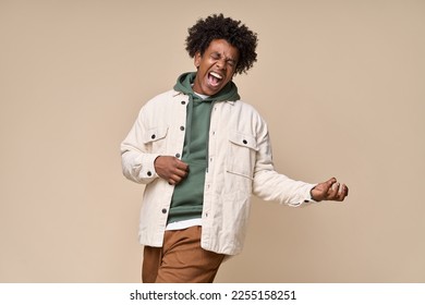 Crazy cool hipster African American guy pretending playing guitar having fun isolated on beige background. Funny funky ethnic generation z teenager fashion model screaming singing and dancing.