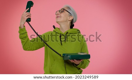 Crazy elderly granny gray-haired woman in casual green hoodie talking on wired vintage telephone of 80s, fooling making silly faces. Senior old grandmother isolated on pink background. People emotions