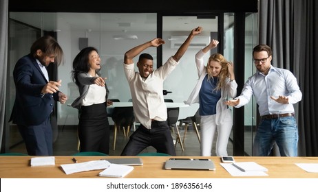 Crazy emotional young african american man having fun with happy diverse mixed race colleagues, dancing together in modern office, celebrating corporate success or profitable goal deal achievement.