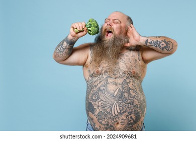 Crazy Fat Pudge Obese Chubby Overweight Man Has Tattooed Big Belly Hold Eat Fresh Vegetable Broccoli Put Hand On Head Sing Song Isolated On Blue Background. Weight Loss Obesity Healthy Diet Concept