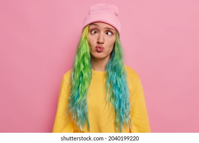 Crazy Female Student With Colored Hair Has Fun Alone Awkward Expression Foolishes Around After All Day Ofstudying Crosses Eyes Pouts Lips Wears Hat And Jumper Poses Against Pink Studio Background.