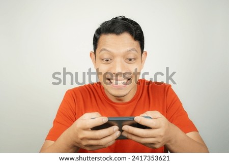 Crazy, funny and excited face of Asian man in orange t-shirt addicted to play mobile game