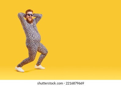 Crazy Funny Fat Man With Big Belly In Pajama Suit With Leopard Print Is Dancing And Fooling Around. Cheerful Chubby Bearded Freak Man In Sunglasses Laughing Loudly On Orange Background. Full Length.