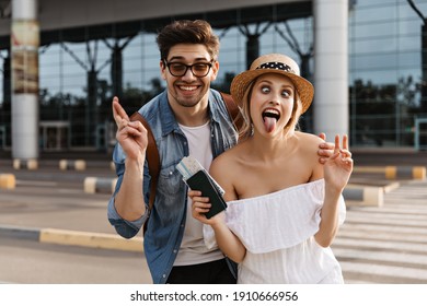 Crazy happy blonde woman in hat and man in sunglasses make funny faces, cross fingers and show v-sign. Cool young girl holds passport near airport.