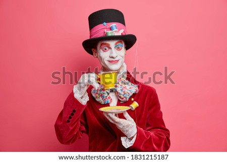 Crazy hatter with professional vivid colorful halloween makeup dressed in costume poses with cup of tea against pink studio background. Gorgeous male character prepares for carnival party. Face art