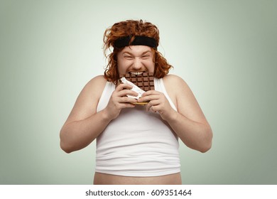 Crazy and hungry overweight young redhead European male with good appetite wearing black sports bra and white undersized tank top biting bar of chocolate, feeling hungry after intensive cardio workout