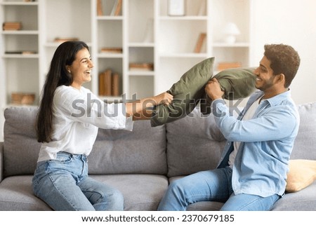 Crazy indian man and woman sitting on couch, having pillow fight and laughing, happy hindu spouses having fun together at home on weekend, copy space