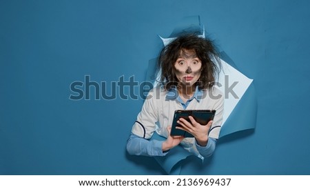 Crazy insane chemist with messy hair using digital tablet and browsing internet, feeling mad funny and doing goofy expressions. Foolish amusing female scientist having wacky grimace.