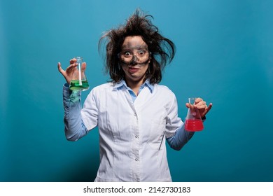 Crazy looking chemist holding Erlenmeyer glass jars filled with unknown chemical compounds while on blue background. Lunatic funny scientist specialist holding flasks filled with liquid substances.