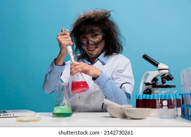 Crazy looking mad chemist grinning creepy at camera while using dropper to to laboratory work after chemical explosion. Foolish silly female lab worker with pipette sitting at desk on blue background.