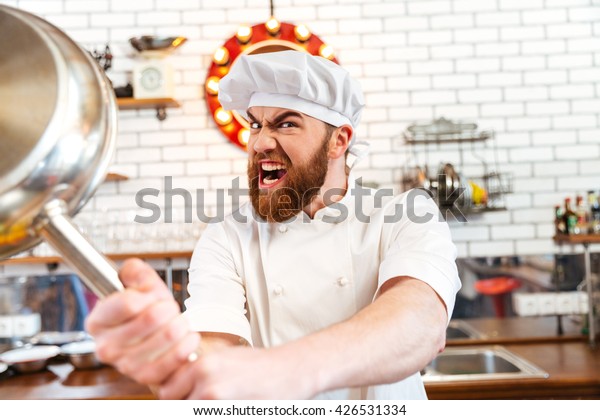Crazy mad chef cook threatening with frying pan\
on the kitchen