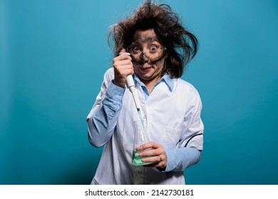 Crazy Mad Scientist Using Pipette To Drip Chemical Compound Inside Glass Beaker Filled With Liquid Toxic Substance. Wacky Chemist With Pipette And Chemistry Flask On Blue Background. Studio Shot.