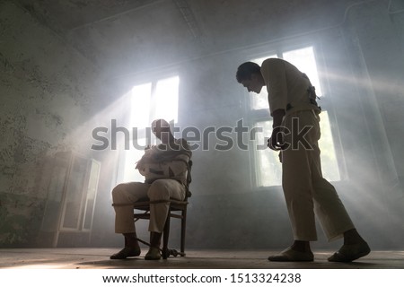 A crazy man in a straitjacket is tied to a chair in an abandoned old clinic and the other insane man coming closer with interest