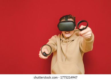 Crazy Man In VR Helmet Plays Video On Controllers Against Red Background, Looks Into The Camera With A Nervous Face. Mad Guy Playing Virtual Reality Games Using A Vr Helmet.