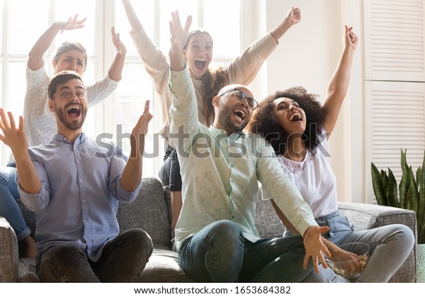 Crazy multiracial people shouting with joy\
celebrating unbelievable victory of favourite sport team. Diverse\
friends gathered together in living room eating pop corn watch\
amusing tv show game\
concept