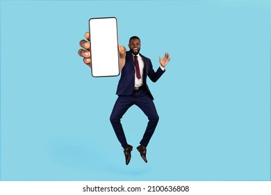 Crazy Offer. Excited Black Businessman Jumping With Blank Smartphone In Hand, African American Male In Suit Showing White Phone Screen At Camera, Recommending Mobile App, Blue Background, Mockup