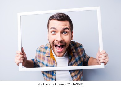 Crazy picture. Handsome young man in shirt holding picture frame in front of his face and smiling while standing against grey background 