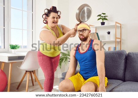 Crazy plus size housewife having fun and hitting her husband with frying pan. Happy, funny, fat woman in hair curlers wants to fight and hits man on head with heavy skillet