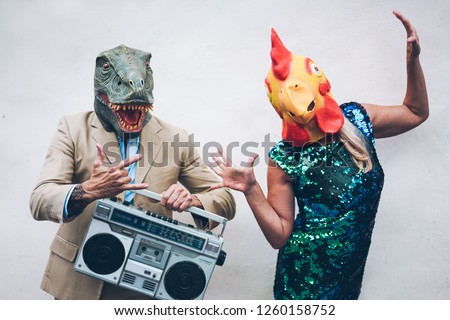 Crazy senior couple dancing for new year's eve party wearing t-rex and chicken mask - Old trendy people having fun listening music with boombox stereo - Absurd and funny trend concept - Focus on faces