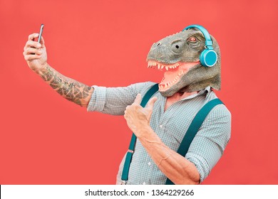 Crazy senior man wearing t-rex mask and taking selfie with mobile smartphone - Hipster older male having fun listening music and dancing outdoor -  Absurd, funny and surreal concepts - Powered by Shutterstock