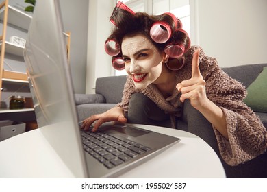 Crazy Smiling Mad Housewife In Cosmetic Face Mask, Hair Rollers, Bathrobe Font Of Laptop Screen With Raised Finger. Funny Cheerful Young Caucasian Woman Having Idea Plotting Something With Sly Smile