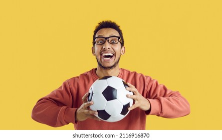 Crazy soccer fan supporting favorite team. Portrait of happy funny excited cheerful black man standing isolated on solid yellow colour background, holding football, looking at camera and smiling