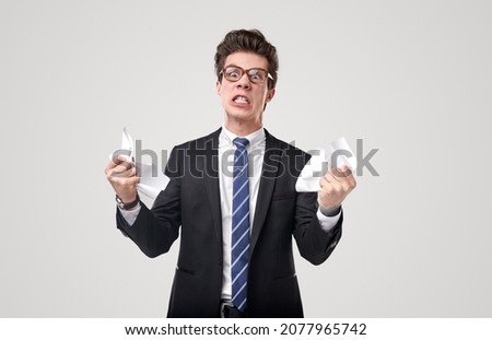 Crazy stressful male executive manager in formal suit and nerd eyeglasses tearing paper document with anger while standing against gray background