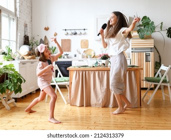 Crazy Weekend With Family. Young Woman Mother With Happy Cute Little Child Daughter In Casual Wear Holding Hair Brushes As Microphone, Singing Karaoke And Dancing In The Modern Cozy Kitchen At Home