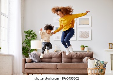 Crazy Weekend With Mom. Joyful Afro American Family Mother And Little Son Holding Hands And Jumping On Sofa In Living Room At Home, Full Length Shot. Mom And Child In Casual Wear Playing Together